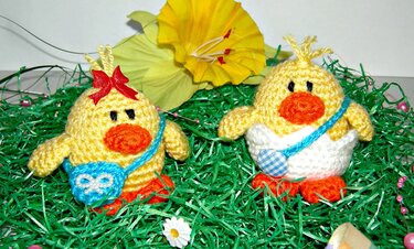 Zwei gelbe gehäkelte Küken mit orangen Schnäbeln und kleinen blauen Täschchen im grünen Ostergras