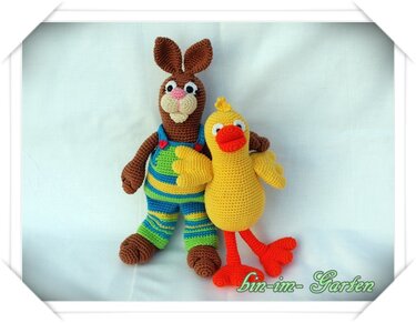 Gehäkelter brauner Hase in blau‑grüner Latzhose neben einer gelben Ente mit orangefarbenem Schnabel und Füßen vor hellem Hintergrund.