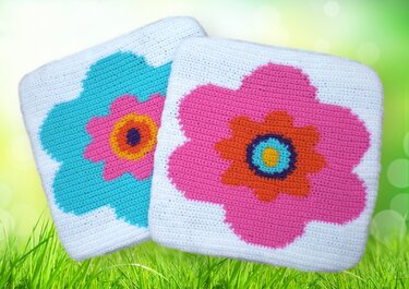 Zwei gehäkelte quadratische Kissenbezüge mit bunten Blumenmotiven auf weißem Grund, auf grünem Gras liegend.