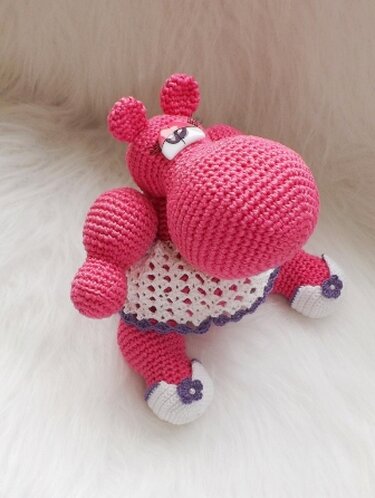 Rosa gehäkeltes Amigurumi-Nilpferd mit weißem Rock und lila Details, sitzend auf hellem Fell