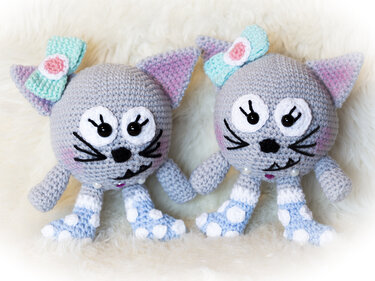 Zwei gehäkelte graue Amigurumi-Katzen mit türkisfarbenen Schleifen und gepunkteten Beinen auf hellem Hintergrund.