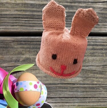 Gestrickter Hasen-Eierwärmer in Rosa neben braunem Ei mit gepunktetem Band auf Holz