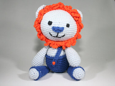 Gehäkelter Amigurumi-Löwe mit orangefarbener Mähne und blauer Latzhose, sitzend