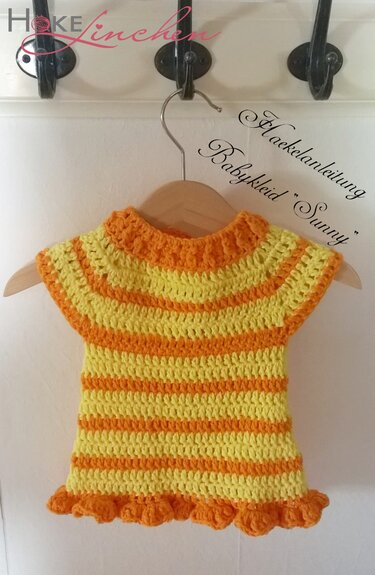 Gestreiftes, gehäkeltes Babykleid in Gelb und Orange an einem Holzkleiderbügel vor weißer Wand.