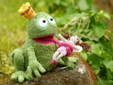 Hand-knit green frog amigurumi wearing a yellow beaded crown holding a small pink-and-white fairy doll on a mossy rock with green foliage behind.