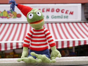 Front-facing knitted frog amigurumi wearing red-and-white striped sweater, blue shorts and red tasselled hat