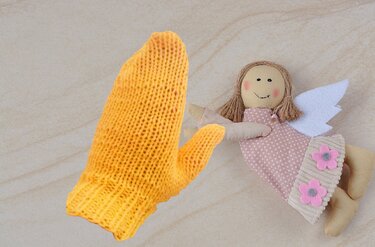 Gelber gestrickter Kinderfäustling neben einer Stoffpuppe mit rosa Kleid und weißen Flügeln auf hellem Hintergrund.