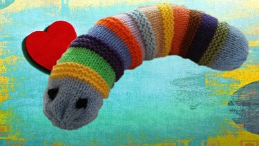 Gestrickte mehrfarbige Raupe mit schwarzen Augen und rotem Herz im Hintergrund