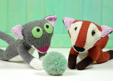 Two knitted amigurumi animals - a gray cat and a red striped fox - playing with a mint-green pom-pom