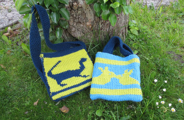 Zwei gestrickte Taschen auf grünem Gras: eine dunkelblau-gelbe Umhängetasche mit Dinosaurier, eine hellblau-gelbe Tragetasche mit Kaninchenmuster.