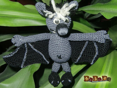Grau-schwarze gehäkelte Fledermaus-Amigurumi mit weißen Haarbüscheln und Wackelaugen vor grünen Blättern
