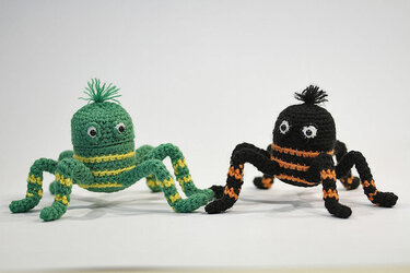 Zwei gehäkelte Amigurumi‑Spinnen, eine grün-gelb gestreift, die andere schwarz-orange gestreift, auf hellem Hintergrund.