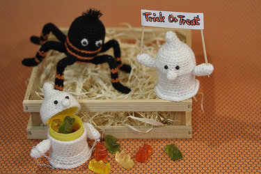 Gehäkelte Halloween-Figuren: schwarze Spinne und zwei weiße Geister; ein Geist hält ein „Trick or Treat“-Schild, ein anderer ist als Süßigkeitenbehälter geöffnet.