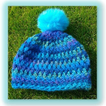 Gehäkelte blau-türkise Mütze mit flauschigem Bommel auf Rasen