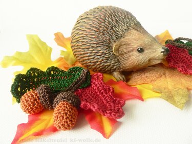 Igel-Dekofigur mit gehäkelten roten und grünen Blättern sowie gehäkelten Eicheln auf herbstlichem Laub