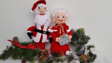 Gehäkelte Amigurumi-Puppen: Weihnachtsmann und Frau Claus mit Geschenk, arrangiert mit Tannenzweigen und Zapfen.