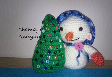 Gehäkelter weißer Schneemann mit blauer Kapuze und rosa Knopf neben grünem gehäkeltem Weihnachtsbaum mit bunten Pailletten