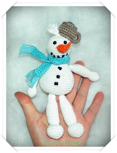 Hand hält kleinen gehäkelten Schneemann mit türkisfarbenem Schal, oranger Nase und brauner Mütze