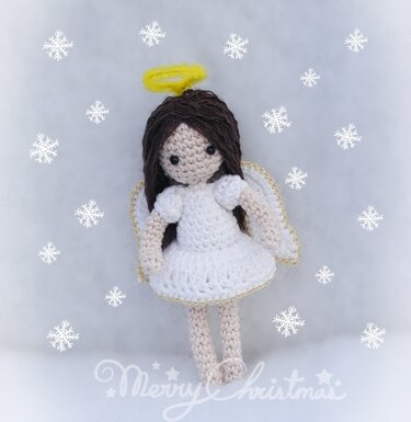 Gehäkelter Amigurumi-Engel mit weißem Kleid, braunen Haaren, gelbem Heiligenschein und goldumrandeten Flügeln vor Schneeflocken-Hintergrund