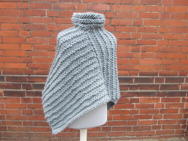 Hellgrauer, grob gestrickter asymmetrischer Poncho mit Rollkragen auf einer Schaufensterpuppe vor roter Backsteinwand