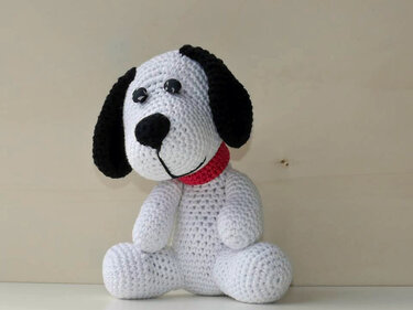 Handgearbeiteter Amigurumi-Hund, weiß mit schwarzen Ohren und rotem Halsband, sitzt auf hellem Untergrund