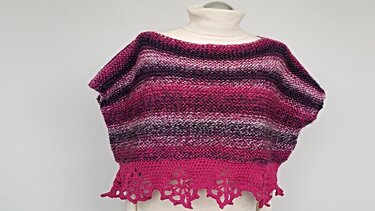 Gestrickter Damenponcho in Fuchsia, Violett und Weiß mit breiten horizontalen Streifen und gehäkelter Lochmusterborte auf einer Schaufensterpuppe.