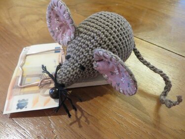 Gehäkelte grau-braune Amigurumi‑Maus mit rosa Stoffohren auf einer Holz‑Mausefalle, darunter ein Geldschein