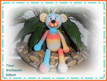 Mehrfarbiger gehäkelter Amigurumi-Teddy mit langen Beinen, sitzend in einer Holzschale vor einer Pflanze