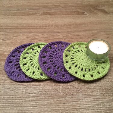 Vier runde, gehäkelte Untersetzer in Lila und Hellgrün auf einem Holztisch, einer davon mit einem Teelicht im Glas.