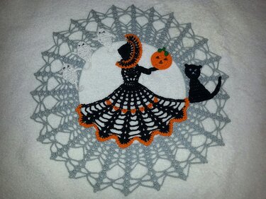 Round crocheted doily with grey lace border, black silhouette holding an orange pumpkin, three white ghost motifs and a black cat applique