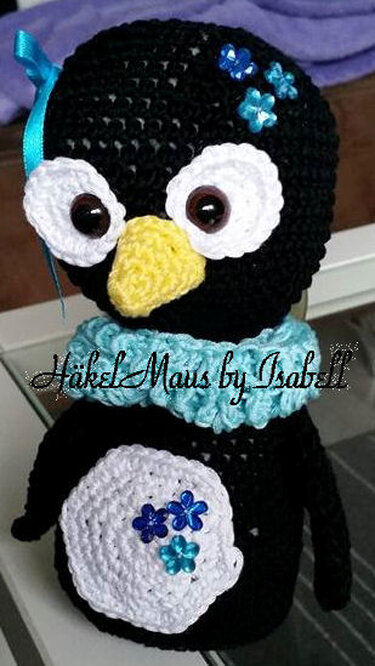 Schwarz-weißer Pinguin-Amigurumi mit gelbem Schnabel, hellblauem Rüschenkragen und blauen Blumenapplikationen