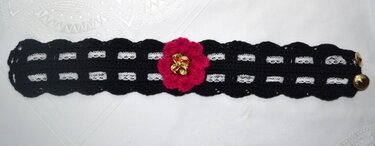 Schwarzes gehäkeltes Armband mit weißem Band, pinker Blüte und goldenen Knöpfen