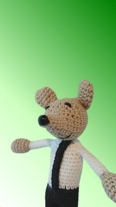 Gehäkelte Amigurumi-Maus mit beigem Kopf, weißem Oberteil und schwarzer Krawatte vor grünem Hintergrund