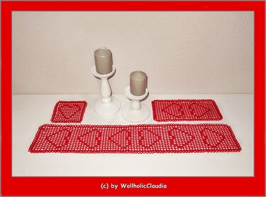 Rotes gehäkeltes Set mit langem Tischläufer, rechteckigem Deckchen und quadratischem Untersetzer mit durchbrochenen Herzmotiven auf weißer Fläche neben zwei weißen Kerzenhaltern mit grauen Kerzen.