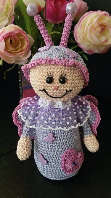 Gehäkelte Amigurumi-Schmetterlingspuppe in Lila mit Perlenfühlern und gepunktetem Stoffkragen vor Blumen