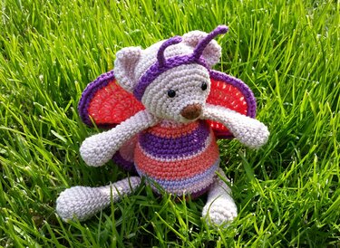 Gehäkelter Amigurumi-Teddybär mit Schmetterlingsflügeln und Fühlern sitzt im grünen Gras