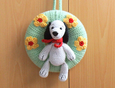 Gehäkelter mintgrüner Türkranz mit gelben Blumen und sitzendem weißen Amigurumi‑Hund mit rotem Schal, an Holztür hängend