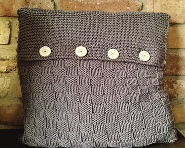 Grau-lila gestrickter Kissenbezug mit vier hellen Knöpfen vor einer Ziegelwand.