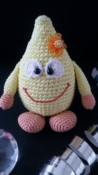 Gehäkelte gelbe Birnenfigur mit Augen, rotem Lächeln, rosa Füßen und orangefarbener Blume