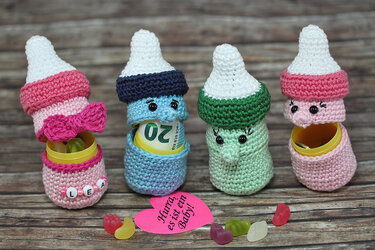 Vier gehäkelte Babyflaschen-Amigurumi in Rosa, Blau und Grün, teils geöffnet als Geschenkverpackung mit Geldschein und gelber Kapsel