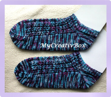Zwei gestrickte Kurzsocken in Blau, Türkis und Violett mit strukturiertem Muster auf hellem Tuch.