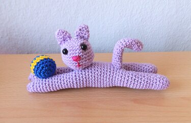 Lavendelfarbene gehäkelte Katze liegend auf Holztisch mit blau-gelbem Ball