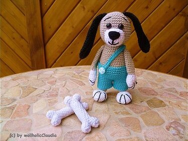 Gehäkelter beiger Amigurumi-Hund mit schwarzen Ohren und türkiser Latzhose auf einem Mosaiktisch; zwei weiße Häkelknochen davor, Holzwand im Hintergrund.