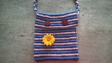 Gehäkelte Henkeltasche mit blau-rosa-grauen Streifen, Klappe, zwei roten Knöpfen und gelber Blume auf grauem, strukturiertem Untergrund.