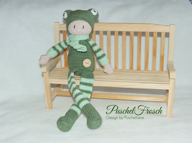 Nahansicht einer gehäkelten Frosch-Puppe mit gestreiften Beinen und Schal auf einer Holzbank