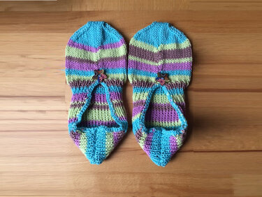 Gestrickte Damenpantoffeln mit bunten Streifen und kleinem blumenförmigem Knopf auf Holzboden.