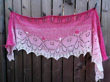 Dreieckstuch gestrickt in Pink und Weiß mit Blatt‑Lace, gezacktem Spitzensaum und kleinen Blütenapplikationen, an einer Holzwand aufgehängt