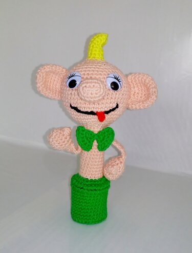 Gehäkelte Amigurumi-Figur mit Wackelaugen, gelber Haarlocke und grüner Fliege vor weißem Hintergrund
