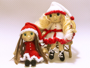 Zwei gehäkelte Amigurumi-Weihnachtspuppen mit Zipfelmützen, roten und weißen Kleidern, langen Haaren; eine sitzt auf einem Korbstuhl.