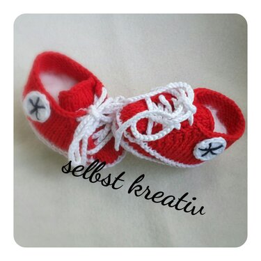 Rote handgestrickte Babyschuhe im Sneaker‑Stil mit weißen Schnürsenkeln auf hellem Untergrund